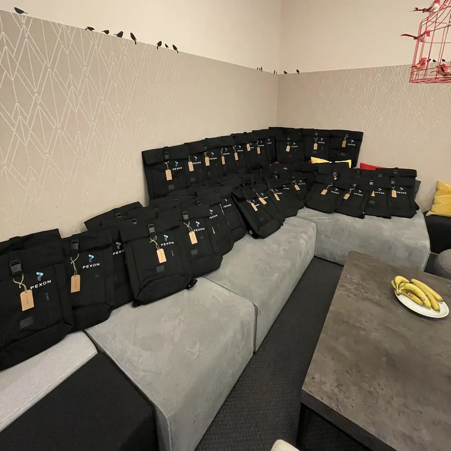 Rund 40 schwarze Rolltop-Rucksäcke mit Pexon-Logo auf einem Sofa